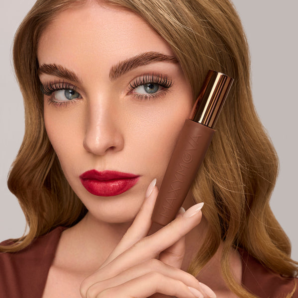 A close-up of a woman with blonde hair holding a brown cosmetic tube near her face against a light gray background.